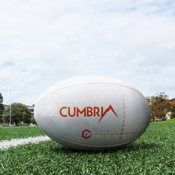 A white rugby ball with an orange logo on a grass field under a cloudy sky.