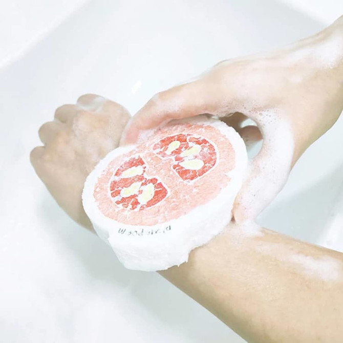 A pink and white expandable dishwashing sponge held in soapy hands over a sink.