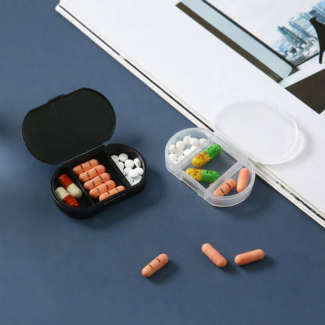 A black pill box with orange and white pills, alongside a clear box containing green and yellow capsules on a dark surface.