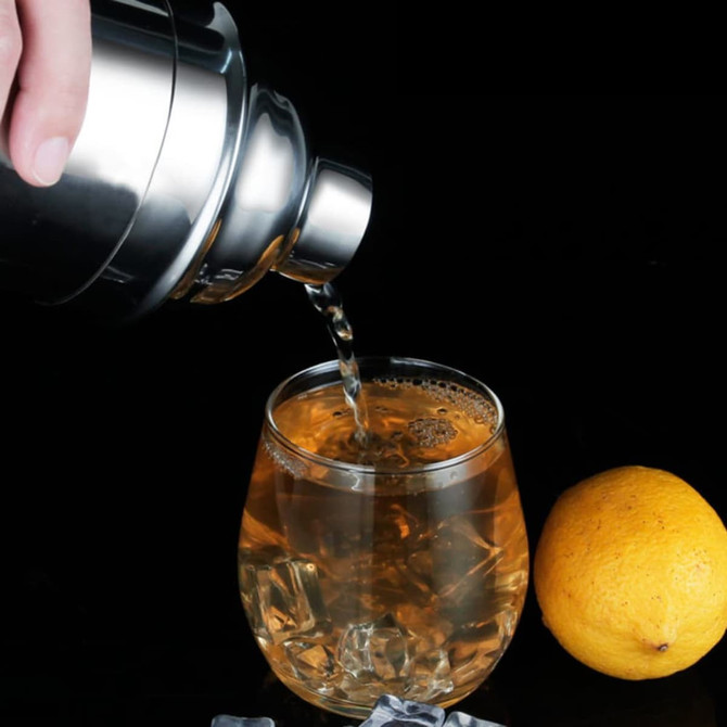 A drink bottle in metallic finish pouring a cocktail into a glass with ice, accompanied by a lemon on the side.
