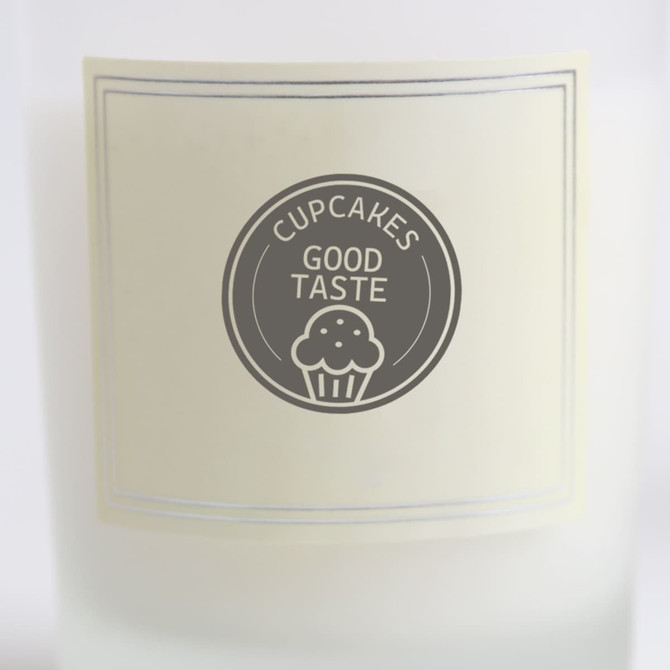 A 2.2oz glass candle featuring a cream-coloured label with a logo and text related to cupcakes and good taste.