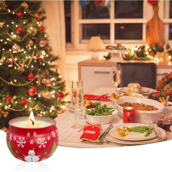 A red tin candle with a snowman design, placed near a festive dinner setting and Christmas tree.