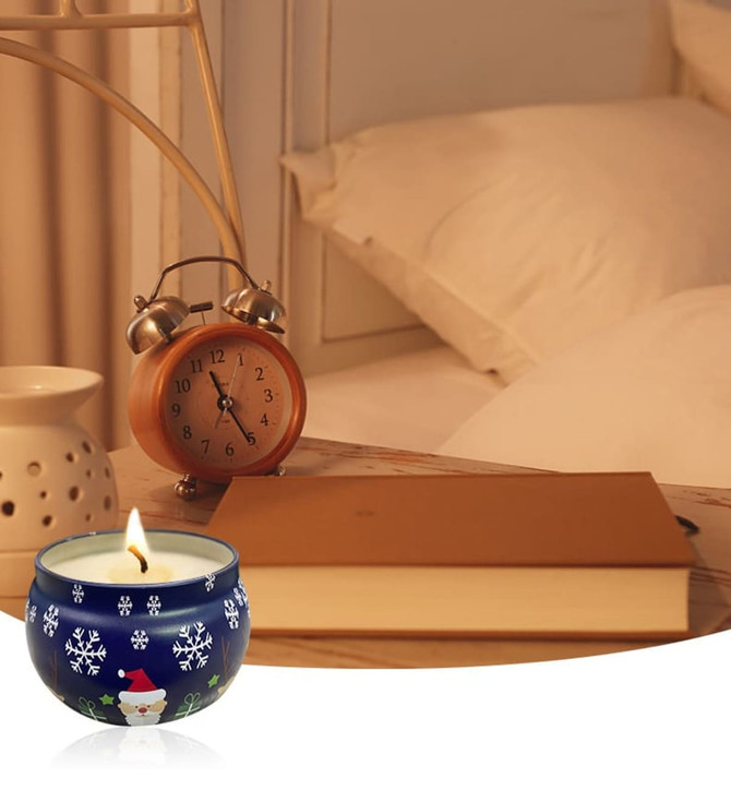 A blue tin candle with a holiday design, surrounded by a bedside table with a clock and a book.