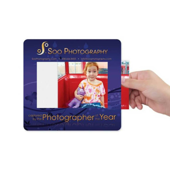 A hand holds a framed mat featuring a colourful design, with an empty space for a photo. It includes a logo.