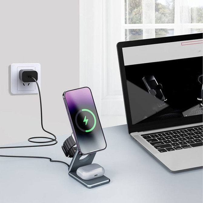 A foldable wireless charger stand in grey, charging a phone and earbuds, placed next to a laptop.