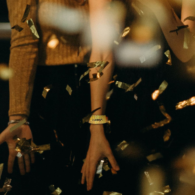 A holographic gold PVC wristband is worn on a wrist, surrounded by golden confetti. The image has a blurred background.