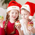 Two children wearing red and white plush Santa hats hold decorated cupcakes, smiling at each other. The hats have a logo.
