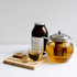 A glass teapot containing yellow tea, a clear cup of green tea, a jar of tea blend, and a wooden serving board.