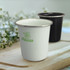 A drink tumbler in white with a green logo and a black tumbler, both with metallic rims, set against a blurred background.