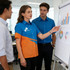 A woman in an orange and blue shirt stands beside a whiteboard, discussing graphs with two men in blue shirts.