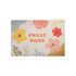 A floral plush logo mat in pastel colours featuring various flowers and the phrase "SWEET HOME."