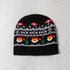 A black knitted beanie featuring festive designs, including smiling Santa faces and snowflakes. It has a logo.