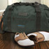 A dark green duffle bag with dark straps, accompanied by brown and white shoes, resting on a wooden floor.