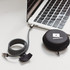 A charging cable in black and grey, coiled next to a round black case with a logo, connected to a laptop.