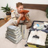 A set of grey travel packing cubes arranged beside a suitcase, with a man sitting on a bed, holding an orange item.