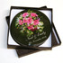 A round, black coaster featuring a floral design with pink roses, presented in a square box.