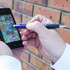 A hand holding a blue and black stylus pen, poised over a smartphone against a brick wall background.