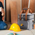 A grey 10L cooler bag with a logo, placed beside a yellow hard hat and a person holding a drink.