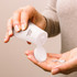 A white sunscreen tube being squeezed, dispensing a small amount of white cream onto a hand. The tube features a logo.