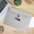 A grey felt laptop bag with a button closure and a logo, placed on a wooden desk alongside stationery and a laptop.