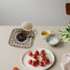 A medium quilted pot holder in a checkered pattern with a logo, placed on a table with a mug and fresh tomatoes.