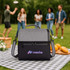 A two-compartment lunch bag in grey and black with a logo, resting on a picnic blanket. People are enjoying a picnic in the background.