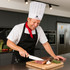 A chef in a white disposable non-woven hat is slicing meat on a wooden cutting board in a modern kitchen.
