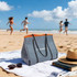 A large grey neoprene beach tote bag with orange accents sits on a towel beside sunglasses and a straw hat on the sand.
