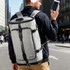 A grey convertible bucket backpack duffel bag with black straps, held by a man in an urban setting, listening to music.