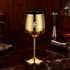 A stainless steel wine goblet in a shiny gold finish, featuring a logo, set on a marble table with bar accessories.