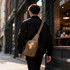 A tactical sling bag in tan is worn crossbody by a person in a black outfit, walking past a café.
