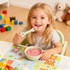 A child smiles while holding bear-shaped green silicone cutlery, enjoying a bowl of pink yoghurt. Colourful toys are in the background.