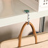A round foldable desk hook in silver, attached to a shelf, with a tan handbag hanging from it.