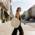 A woman walks along a street carrying a cream mesh grocery bag, featuring an inner section and a logo.