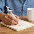 A white acrylic pen with a decorative clip is being used to write on a lined notebook beside a cup.