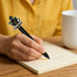 A black acrylic pen with a cartoon cat design is held over a notepad and a white cup on a wooden surface.