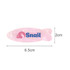 A fish-shaped iridescent hair clip in pink with a logo. Dimensions are 6.5 cm long and 2 cm wide.