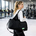 A black duffle bag with a strap, carried by a woman in a gym setting. It features a logo on the side.