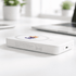 A white wireless charging power bank with a logo, resting on a desk near plants and a laptop.