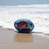 A blue and red soft AFL football rests on wet sand near the ocean, with waves gently rolling in.