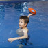 A child in a swimming pool holds an orange soft rugby ball, preparing to throw it. The water is blue and clear.
