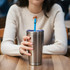 A drink bottle with a silver finish features a blue straw topped with a brown brim hat, held by a person in a light jumper.