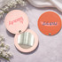 A round makeup mirror with a light pink exterior and a vibrant orange exterior, featuring logos on each side.