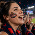 A woman with a temporary tattoo on her cheek, smiling passionately at a sports event, surrounded by excited fans.