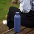 A drink bottle in matte navy blue, featuring a sleek design and a logo, resting on a wooden bench.