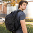 A black backpack is worn by a young man, featuring multiple pockets and a sturdy design, outdoors.