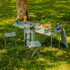 A cooler box in grey with a foldable table and two small chairs, surrounded by cans and snacks on a grassy area.
