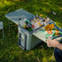 A green cooler box with a table and chair, filled with drinks and snacks, set on grass.