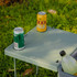 A light green table with two cans and a drink bottle on it, situated on grassy ground. The table has a logo.