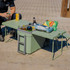 A green cooler box with an attached table and chair, holding drinks and snacks on a paved surface.